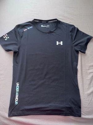 Svart reflektiv under armour t shirt - Svart kortärmad träningströja från Under Armour med tryckt logga på bröstet och text längs sidan. Tillverkad i ett lätt och snabbtorkande funktionsmaterial, perfekt för löpning eller gym. Rund halsringning och diskreta detaljer på ärmen.