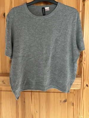 Grå stickad t-shirt från Selected Femme - En enkel och stilren grå stickad t-shirt från Selected Femme. Modellen har korta ärmar, rund halsringning och ribbade kanter. Perfekt för lager på lager och funkar året runt. Materialet känns mjukt och lätt.