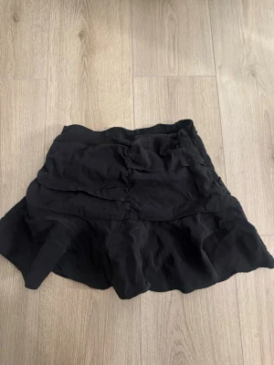 Svart volangkjol från Zara - Kort svart kjol från Zara med volangdetaljer och lätt rynkad design. Kjolen har en flowy passform och är tillverkad i ett mjukt syntetmaterial som ger en skön känsla. Har även inbyggda shorts inuti