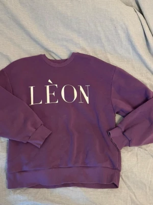 Lila sweatshirt med LÈON-tryck - En lila sweatshirt med stort vitt LÈON-tryck på bröstet. Tröjan har rund halsringning, ribbade muddar vid ärmslut och nederkant samt en avslappnad passform. Perfekt för dig som gillar en enkel men snygg stil.