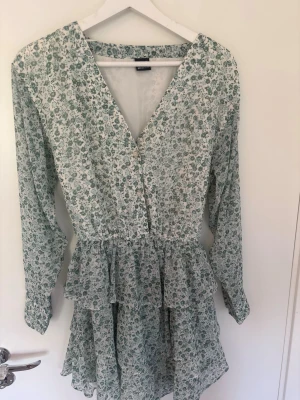 Grön blommig volangklänning från Gina Tricot  - Säljer en supersöt klänning med grönt blommönster, v-ringning och knappar framtill. Klänningen har långa ärmar och volangdetaljer i kjolen. Klänningen är i storlek xs men passar även s då den är stor i storleken. Knappt användt så den är i jättebra skick. 