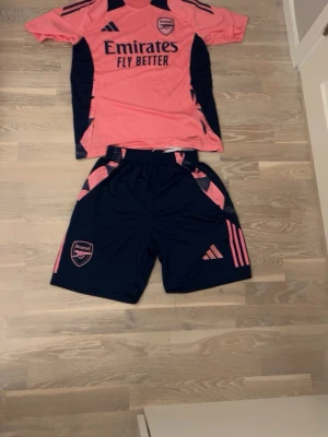 Arsenal träningsset rosa/marinblå - Säljer ett Arsenal träningsset från Adidas i rosa och marinblått. Setet består av en kortärmad tröja med Arsenal-logga och Emirates-tryck samt matchande shorts med elastisk midja och fickor. Perfekt för fotbollsträning eller gymmet.
