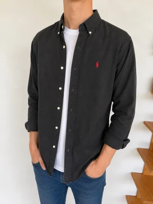 Svart skjorta från Ralph Lauren - Klassisk svart skjorta från Ralph Lauren med knappslå och vit knappdetalj. Skjortan har lång ärm och den ikoniska röda loggan broderad på bröstet. Tillverkad i mjuk bomull och har en normal passform. Perfekt till jeans eller chinos.