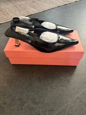 Svarta klackskor  - Stilrena svarta ”kitten heels” i lack med öppen häl. Skorna har en elegant, smal klack i metallic och justerbar rem med spänne. Perfekta för dig som vill ha en modern och trendig look. Materialet är glansigt och ger en lyxig känsla. Helt nya/oanvända och säljs då de är för stora💞