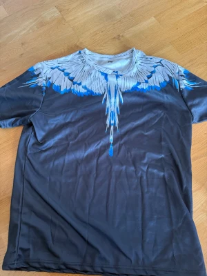 Svart t-shirt med blått fjädermönster - Snygg svart t-shirt med ett unikt blått och vitt fjädermönster runt halsen och axlarna. T-shirten har rund hals och korta ärmar. Materialet känns mjukt och lätt, perfekt för en cool och avslappnad stil.