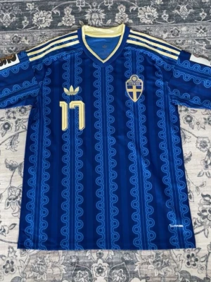 Blå Sverige fotbollströja nr 17 - Säljer en blå fotbollströja med Sveriges landslagsemblem och nummer 17 i gult. Tröjan har ett unikt mönster, V-ringning med gul och blå kant samt gula ränder på axlarna. Tillverkad i ett lätt och ventilerande material, perfekt för träning eller match.