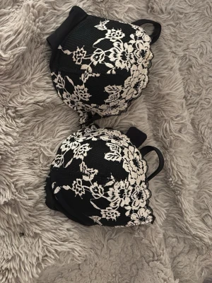 Svart push-up bh med vit spets - Snygg svart push-up bh med vadderade kupor och dekorativ vit spets i blommönster på framsidan. Axelbanden är justerbara och bh:n har hakknäppning bak. Materialet är en mix av spets och slät textil för en lyxig känsla.