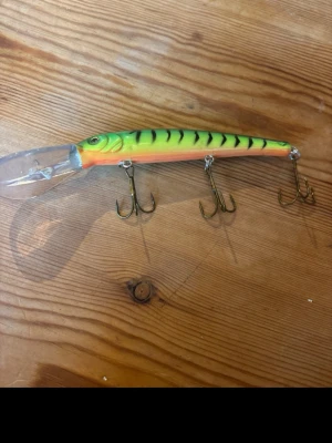 Grön-orange fiskedrag med trekrokar - Säljer ett färgglatt fiskedrag i grönt och orange med svarta ränder och tre trekrokar. Draget har en långsmal form och en genomskinlig sked framtill för djupare fiske. Perfekt för dig som gillar att fiska gädda eller abborre.