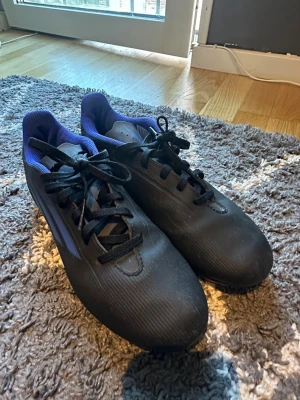 Svarta fotbollsskor med blå detaljer - Säljer ett par stilrena fotbollsskor i svart med blå insida och svarta snören. Skorna har en låg profil och är tillverkade i syntetmaterial för bra bollkontroll. Perfekta för dig som vill ha en klassisk look på planen.