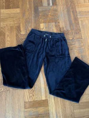 Mörkblå velour mjukisbyxor - Mörkblå mjukisbyxor i mjuk velour med bred resår i midjan och dragsko. Byxorna har två fickor fram och två bakfickor med knappdetaljer. Perfekta för chill dagar och riktigt sköna att ha på sig. OBS: litet hål i skrevet 