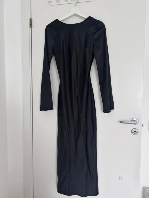 Svart långklänning med knyt framtill - Snygg svart långklänning med lång ärm och djup v-ringning framtill. Klänningen har en trendig knyt-detalj vid bysten och är figurnära i modellen. Perfekt för dig som vill ha en stilren och elegant look.