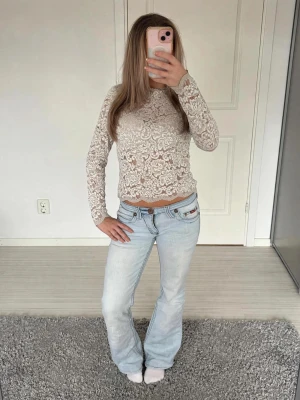 Beige spetsblus med lång ärm - Säljer en beige, transparent spetsblus med lång ärm och rund hals. Blusen har ett detaljerat blommönster i spets och är figurnära i modellen. Perfekt för dig som gillar romantisk och trendig stil.
