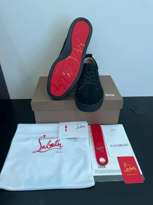 Svarta Christian louboutins  - Snygga svarta sneakers från Christian Louboutin med ikonisk röd sula och klassisk låg design. Skorna har ovandel i mocka och detaljer i svart läder. Perfekta för dig som vill sticka ut med exklusiv stil och streetkänsla.
