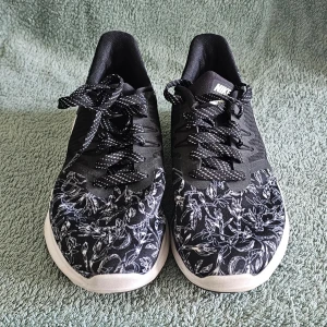 Svarta Nike sneakers med blommönster - Snygga svarta Nike sneakers med vitt blommönster på tå och sidor. Skorna har vita sulor, svart/vita skosnören och den klassiska vita Nike-loggan på sidan. Ovandelen är i textilmaterial som andas och ger en sportig look.
