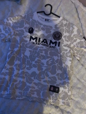 Inter Miami x BAPE camo t-shirt limited - Snygg t-shirt i samarbete mellan Inter Miami och A Bathing Ape. Tröjan har ett gråvitt camomönster, svart tryck med MIAMI och BAPE-logga samt Inter Miami-emblem på bröstet. Perfekt för dig som gillar streetwear och fotboll.