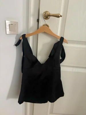 Svart satinlinne med knytband - Stilrent svart linne i satin med breda axelband som knyts upptill. Toppen har en lätt glansig finish och en figurnära passform. Perfekt att styla med jeans eller kjol för en trendig look.
