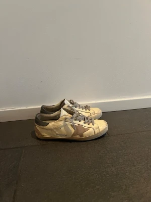 Golden Goose Superstar sneakers - Säljer ett par Golden Goose Superstar sneakers i vitt läder med grå detaljer och en beige stjärna på sidan. Skorna har grå snörning och en mörkgrå hälkappa. Klassisk låg modell med vintage-inspirerad design och coola detaljer. Sulan på en så där fram är det hål i men kanske går att limma ihop!