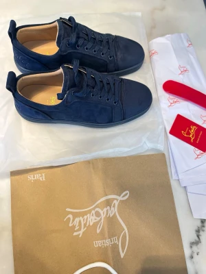 Mörkblå Christian Louboutins  - Snygga mörkblå sneakers i mocka från Christian Louboutin. Skorna har klassisk låg profil, rund tå och matchande mörkblå snörning. Insidan är ljusbeige och sulan är ton-i-ton med ovandelen. Perfekta för dig som gillar stilrena och exklusiva detaljer.