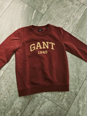 Vinröd sweatshirt från GANT - Säljer en vinröd sweatshirt från GANT med guldtext 'GANT 1949' på bröstet. Tröjan har rund halsringning och ribbade muddar vid ärmslut och nederkant. Perfekt för dig som gillar klassisk college-stil.