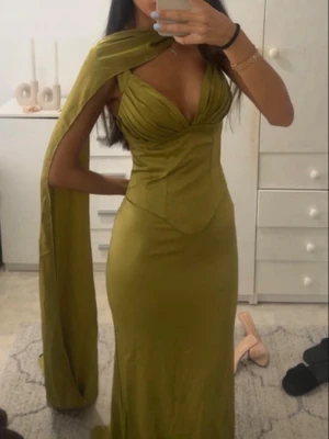 Grön långklänning med cape - Säljer en elegant grön långklänning med djup v-ringning och figurnära passform. Klänningen har en unik cape-detalj som ger en extra dramatisk look. Perfekt för dig som vill sticka ut på bal eller fest.