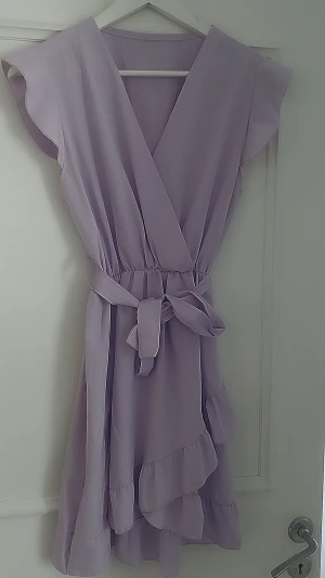 Ljuslila omlottklänning med volang - Superfin ljuslila omlottklänning med volangdetaljer och knyt i midjan. Klänningen har v-ringning, ärmlös design och kort längd. Perfekt för sommarens alla tillfällen!