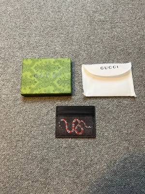 Gucci korthållare med ormprint - Säljer en svart korthållare från Gucci med klassiskt GG-mönster och en cool röd och vit orm på framsidan. Tillbehör som original ask och dustbag medföljer. Perfekt för dig som vill ha något stilrent men ändå sticker ut.