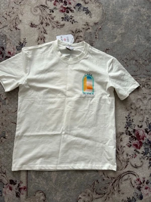 Vit Casa Blanca t-shirt med tryck - Vit t-shirt från Casa Blanca med färgglatt grafiskt tryck på bröstet i blått, orange och rosa toner. Klassisk passform med rund hals och korta ärmar. Tillverkad i mjuk bomull för en skön känsla. Lite bred runt midjan som ger en snygg baggy look.