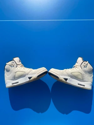 Air Jordan 5 Retro - Säljer ett par vita höga  Air Jordan 5 Retro sneakers med svarta detaljer och meshpaneler på sidorna. Skorna har klassisk snörning, vadderad plös med Jumpman-logga och robust sula. Perfekta för dig som gillar streetwear och sportig stil.