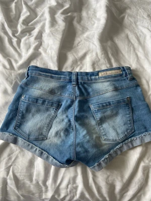 snygga jeansshorts - Säljer ett par klassiska blå jeansshorts med rå kant och femficksmodell. Shortsen har bälteshällor, knapp och dragkedja framtill samt slitna detaljer för en avslappnad look. Perfekta till sommaren och lätta att matcha med olika stilar.