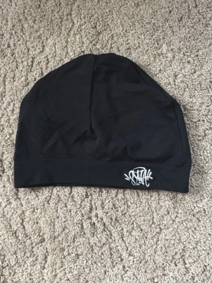 Svart SynaWorld mössa - Svart, slimmad mössa i stretchigt tyg med vit SynaWorld logga framtill. Perfekt för dig som gillar en enkel och stilren look med en touch av streetstyle. Passar bra under jackan eller till vardagsoutfiten. 