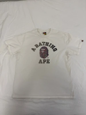 Vit BAPE Tshirt - Säljer en vit t-shirt från A Bathing Ape med det klassiska BAPE-huvudet och texten 'A BATHING APE' i svart och grått framtill. T-shirten har korta ärmar och rund hals. Perfekt för dig som gillar streetwear och vill ha något stilrent men ändå ikoniskt. Den har några små fläckar, med de går lätt bort i tvätten!
