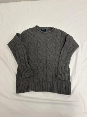 Grå kabelstickad tröja Polo Ralph Lauren - Kabelstickad grå tröja från Polo Ralph Lauren i italienskt garn. Klassisk rund halsringning, ribbade muddar och nederkant. Perfekt för lager på lager och en tidlös look.