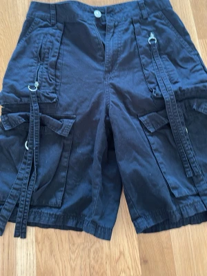 Svarta cargo shorts från H&M Divided - Säljer ett par svarta cargo shorts från H&M Divided med flera fickor, dekorativa band och metallringar. Shortsen har en avslappnad passform och är perfekta för sommaren. Tillverkade i bomull för en skön känsla.