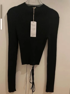 Svart stickad topp med snörning - Trendig svart stickad topp med långa ärmar och snörningsdetalj i ryggen. Kortare modell med rund halsringning och ribbad struktur. Perfekt för att skapa en cool och stilren look.