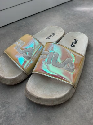 Holografiska Fila sandaler - Säljer ett par snygga Fila sandaler i storlek 39 med holografiskt band i skimrande pastellfärger och stor vit Fila-logga. Sulan är vit och sandalerna har en klassisk slip-in modell. Perfekta för sommaren eller att chilla i hemma.