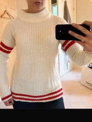 Vit stickad tröja med röda ränder - Säljer en ribbstickad vit tröja med bred polokrage och röda ränder på ärmar och nedtill. Tröjan har lång ärm och en avslappnad passform, perfekt för kalla dagar. Materialet ser ut att vara mjukt och tjockt, troligen ull eller akryl.