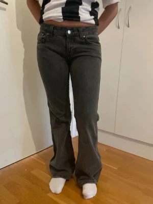 Mörkgrå bootcut jeans - Säljer ett par mörkgrå bootcut jeans med klassisk femficksdesign och knappar på bakfickorna. Jeansen har hög midja och är tillverkade i ett mjukt denimtyg som sitter snyggt över höfterna och låren innan de blir vidare nedtill.