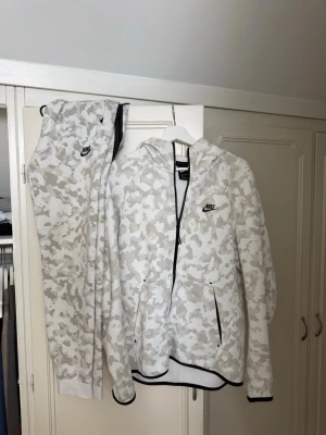Nike tech fleece komplett med vit camomönster - Nike komplett i vitt och ljusgrått camomönster med dragkedja framtill, huva och svarta detaljer vid dragkedja och muddar. Tillverkad i mjukt material och har fickor på sidorna. Perfekt för en avslappnad och sportig stil. Två fläckar finns, bak på byxan och bak på tröjan
