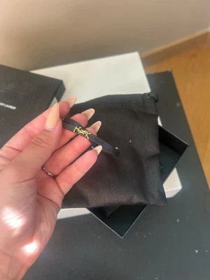 Svart Saint Laurent armband i läder - Snyggt svart armband från Saint Laurent i läder med guldfärgat YSL-logo som detalj. Armbandet har en tunn, minimalistisk design och stängs med ett elegant guldfärgat spänne. Använt endast fåtal gånger!! Nypris 3000kr, pris kan diskuteras 