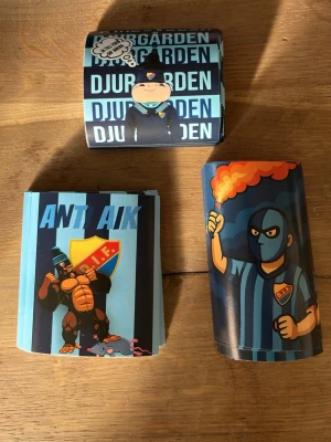 100st Djurgården klistermärken  - Ett gäng klistermärken med Djurgården-tema. Motiven inkluderar en maskerad supporter med bengal, en tecknad figur i DIF-kläder och ett anti-AIK-märke med sköldpadda. Färgerna är främst blå, gul och orange. Perfekt för att dekorera laptop, cykel eller mobil.