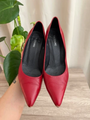 Röda pumps från Vezzano - Snygga röda pumps från Vezzano med spetsig tå och klassisk klack. Skorna är i slätt läder och har en elegant siluett som ger en stilren look. Perfekta för dig som vill sticka ut med färg och stil.