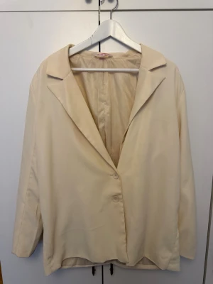 Krämvit oversized kavaj med knappar - Stilren krämvit kavaj i oversized modell med två knappar framtill. Klassisk krage och helfoder ger en clean look. Tillverkad i ett slätt, mjukt tyg som ger en avslappnad men ändå uppklädd känsla. Perfekt att styla lager på lager.