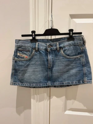 Diesel jeanskjol - Säljer en kort blå diesel jeanskjol i klassisk denim med fem fickor och broderade detaljer på bakfickorna. Kjolen har bälteshällor, knapp och dragkedja framtill samt en liten logga på fickan. Perfekt för dig som gillar en trendig och avslappnad stil. Priset kan diskuteras 