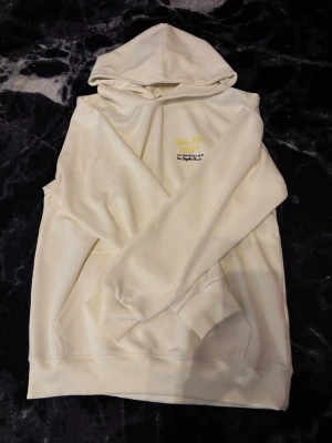 Vit hoodie från Gallery Dept. - Snygg beige hoodie från Gallery Dept. med tryck i gult och svart på bröstet. Klassisk huva och känguruficka framtill. Tillverkad i mjukt material som känns skönt mot huden. Perfekt för dig som gillar streetwear och enkel stil. 