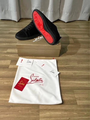 Svarta sneakers från Christian Louboutin - Stilrena svarta sneakers från Christian Louboutin med klassisk röd sula och diskret logga. Skorna har en låg silhuett, svarta skosnören och är tillverkade i mocka och läder. Perfekta för dig som vill ha en lyxig och trendig look.