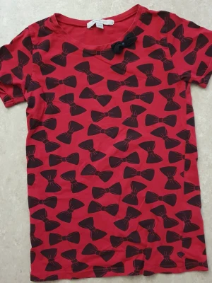 T-shirt - Cool röd t-shirt med svarta rosetter över hela tröjan och en extra tygrosett vid halsen. T-shirten har rund halsringning och korta ärmar. Perfekt för dig som vill sticka ut lite extra med ett lekfullt mönster.