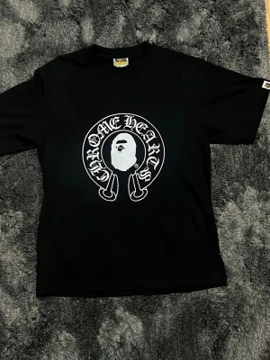 Svart Chrome Hearts x BAPE T-shirt - Säljer en svart t-shirt med Chrome Hearts och BAPE Baby Milo tryck både fram och bak. T-shirten har rund hals, korta ärmar och är gjord i mjuk bomull. Stora grafiska prints i vitt ger en streetwear vibe. Perfekt för dig som gillar unika samarbeten och cool design.