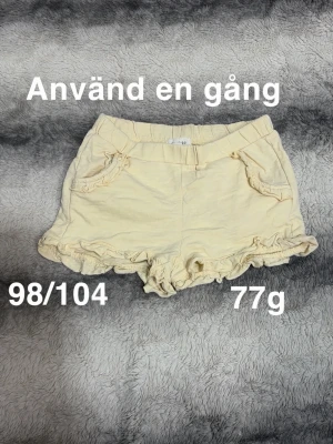 Gula shorts med volangdetaljer - Söta gula shorts med volangkant och dekorativa fickor framtill. Shortsen har elastisk midja och är perfekta för varma dagar. 🌸 Använd en gång. Storlek 98/104