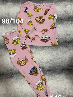 Rosa Paw Patrol pyjamas barn - Söt rosa pyjamas med långärmad tröja och byxor, dekorerad med färgglada Paw Patrol-figurer. perfekt för mysiga kvällar hemma. Elastisk midja på byxorna för extra komfort.🌸 Två svaga fläckar. Storlek 98/104