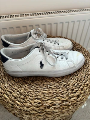 Vita sneakers från Polo Ralph Lauren - Snygga vita sneakers från Polo Ralph Lauren med klassisk låg profil och marinblå logga på sidan. Skorna har vita snören, marinblå detalj vid hälen och är tillverkade i ett tåligt lädermaterial. Perfekta för en clean och sportig look. Som du kan se på bilderna har högra skon en liten defekt framtill. 